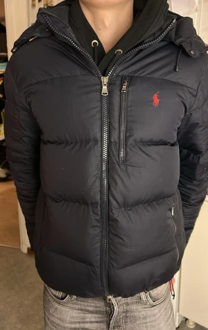 Mörkblå pufferjacka från Ralph Lauren - Snygg mörkblå pufferjacka från Ralph Lauren med röd logga på bröstet. Jackan har huva, dragkedja framtill, bröstficka med dragkedja och två sidofickor. Perfekt för kalla dagar och har en quiltad look. Har en trasig dragkedja och några små hål