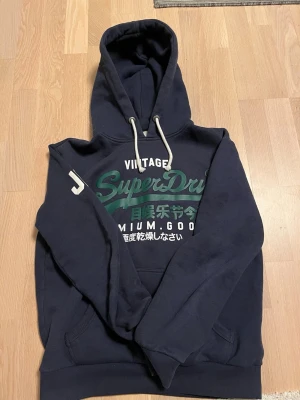 Mörkblå Superdry hoodie med tryck - Mörkblå hoodie från Superdry med stor grön och vit logga samt japansk text på bröstet. Klassisk huva med vita dragsnören och känguruficka framtill. Tryck med 'JPN' på ena ärmen. Perfekt för en avslappnad streetwear-look.