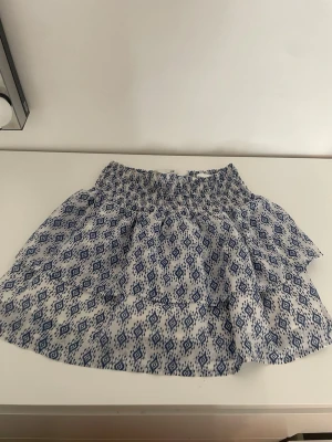 Kortkjol med blått mönster - Säljer en kort kjol med vitt och blått geometriskt mönster. Kjolen har en smockad midja och är lätt och luftig, perfekt för varma dagar. Materialet känns som bomull och kjolen har en volang för extra stil.