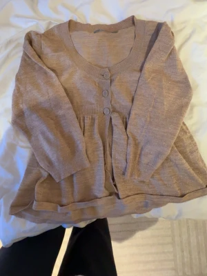 Beige glittrig kofta med knappar - Söt beige kofta med glittriga inslag och tre dekorativa knappar framtill. Står storlek L men passar mer S/M. Koftan har en rund halsringning, lång ärm och lätt utställd passform nedtill. Perfekt för dig som gillar en feminin och mjuk stil.