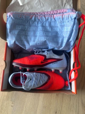 Nike Phantom elite fotbollsskor ”scary good” - Snygga Nike Phantom 6 elite fotbollsskor i färgen ”scary good” rött och blå med svarta detaljer. Med AG platta och med skopåsen som ingår. Endast använda 1 gång. Säljer eftersom de inte passar mig.