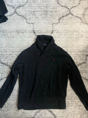 Mörkgrå tröja från Polo Ralph Lauren - Snygg mörkgrå tröja från Polo Ralph Lauren med sjalkrage och två knappar vid halsen. Tröjan har lång ärm och den klassiska röda loggan broderad på bröstet. Perfekt för dig som gillar stilrena och tidlösa plagg.