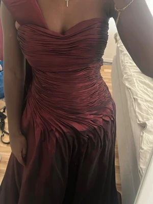 Vinröd långklänning  - Elegant vinröd långklänning med vacker drapering över byst och midja. Klänningen är axelbandslös och har en figurnära passform upptill som övergår i ett svepande kjolparti. Perfekt för bal eller festliga tillfällen.