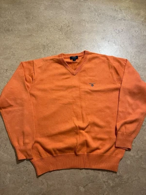 Orange stickad tröja från Gant - Snygg orange stickad tröja från Gant med V-ringning och diskret logga broderad på bröstet. Tröjan har ribbade muddar vid ärmslut och nederkant, vilket ger en klassisk och stilren look. Perfekt att matcha med jeans eller chinos.