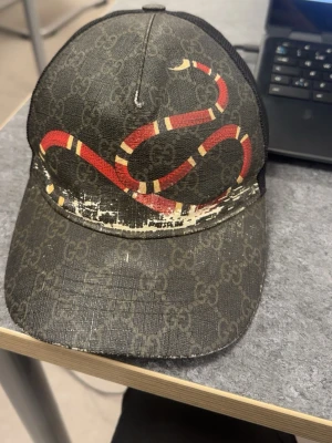 Gucci keps Snake - Snygg keps från Gucci med klassiskt GG-mönster i grått och svart. Framsidan pryds av en röd och vit orm som slingrar sig över kepsen. Baksidan har mesh-tyg för extra ventilation och insidan har en Gucci-etikett. Perfekt för dig som vill sticka ut med en exklusiv accessoar.