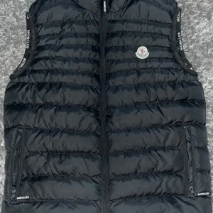 Svart dunväst från Moncler - Snygg svart dunväst från Moncler med quiltad design och dragkedja framtill. Västen har två fickor med dragkedja och Moncler-logga på bröstet samt logotypdetaljer längs ärmöppningarna. Perfekt för lager på lager och streetwear.