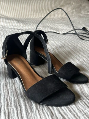 Svarta sandaler med blockklack - Säljer ett par stilrena svarta sandaler med bred rem över tårna och justerbart spänne vid ankeln. Skorna har en bekväm blockklack och ovandelen är i mockaimitation. Perfekta till sommaren och passar till många olika outfits.