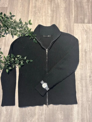 Svart stickad ziptröja 157 - Svart stickad tröja från 157 med dragkedja framtill och hög krage. Tröjan har ribbstickad struktur och långa ärmar. Perfekt för lager på lager och enkel att matcha med olika outfits.