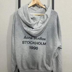 Acne Studios Hoodie ~ Grå - 🔹 Grey hoodie in a relaxed fit with a soft and casual silhouette  🔹 Clean Scandinavian design from Acne Studios with a minimalist aesthetic  🔹 Bold black back print featuring “Acne Studios Stockholm 1996”  🔹 Large centered text graphic that creates a strong streetwear look  🔹 Neutral grey color that pairs easily with many outfits  🔹 Soft cotton sweatshirt fabric designed for comfort and everyday wear  🔹 Adjustable hood that adds a laid-back and practical element