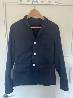 Mörkblå kavaj från Dondup - Stilren mörkblå kavaj från Dondup med tre vita knappar framtill, tre utanpåfickor och klassisk krage. Tillverkad i ett mjukt bomullstyg som ger en avslappnad men ändå dressad look. Perfekt för dig som vill ha en modern och enkel kavaj.