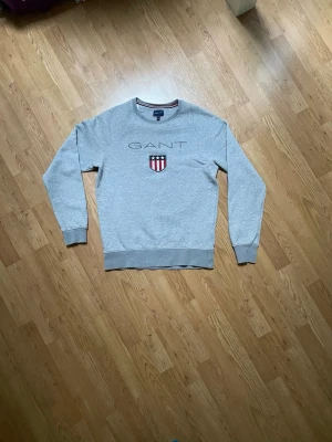 Grå sweatshirt från Gant - Klassisk grå sweatshirt från Gant med rund halsringning och tryckt logga samt sköld på bröstet. Tröjan har långa ärmar och ribbade muddar vid ärmslut och nederkant. Perfekt till en avslappnad stil.
