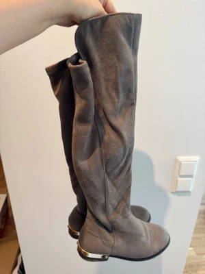 Gråa overknee stövlar med metallhäl - Snygga gråa overknee stövlar i mockaimitation med rund tå och låg klack. Stövlarna har en cool detalj i form av en metallkant på hälen som ger extra edge till looken. Perfekta att styla med kjol eller jeans för en trendig outfit.