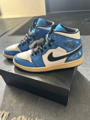 Air Jordan 1 Mid blå och vita sneakers - Snygga Air Jordan 1 Mid sneakers i blått, vitt och svart med klassisk Nike Swoosh på sidan. Skorna har svart snörning, vadderad krage och en beige sula. Tillverkade i läder och syntetmaterial, perfekta för dig som gillar streetwear och ikonisk design.