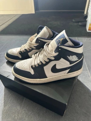 Nike Air Jordan 1 Mid marinblå/vit - Snygga Nike Air Jordan 1 Mid sneakers i marinblått och vitt med klassisk Jordan-logga på sidan och tungan. Skorna har perforerad tå, vita snören och en robust gummisula. Materialet är en mix av läder och textil för både stil och komfort.
