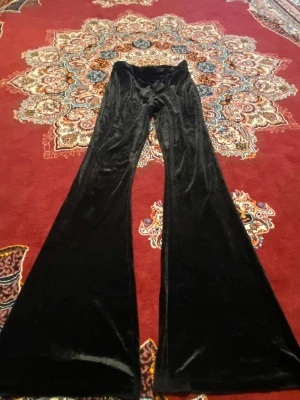 Svarta utsvängda leggings från Kappahl - Säljer ett par svarta leggings från Kappahl i mjuk och glansig sammet. De har en utsvängd passform som ger en cool retrostil och är perfekta för dig som gillar att sticka ut. Materialet är stretchigt och bekvämt, vilket gör dem lätta att bära hela dagen.