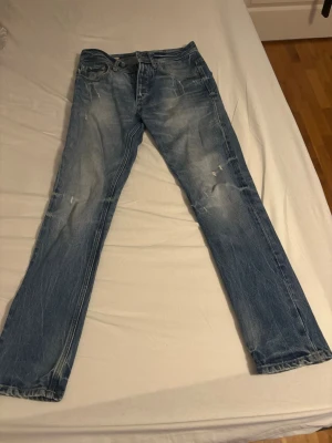 Blå raka jeans med slitningar - Säljer ett par blå jeans med raka ben och snygga slitningar framtill och baktill. Jeansen har klassisk femficksdesign och ljus tvätt för en avslappnad look. Perfekta för dig som gillar en casual stil.