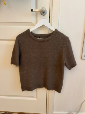 Brun stickad kortärmad tröja - Mysig brun stickad tröja med korta ärmar och rund hals. Tröjan har en enkel, rak passform och ribbade kanter vid ärmslut och nederkant. Perfekt för lager på lager och en avslappnad stil.