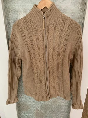 Beige kabelstickad kofta med dragkedja - Snygg beige kabelstickad kofta från Josephine Prandina med hög krage och dragkedja framtill. Koftan har ribbade muddar och ett klassiskt mönster längs hela plagget. Perfekt för lager på lager och en stilren look.