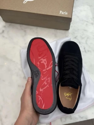 Christian Louboutin svarta sneakers - Snygga svarta sneakers från Christian Louboutin i mocka med klassisk röd sula och svarta skosnören. Ikonisk design med låg profil och exklusiv känsla. Perfekta för dig som vill sticka ut med stilrena detaljer och lyxig finish.
