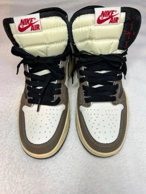 Nike Air Jordan 1 Travis Scott High - Snygga Nike Air Jordan 1 Travis Scott High sneakers i brunt, vitt och svart med ikonisk omvänd swoosh och röda detaljer på plösen. Skorna har högt skaft, svarta skosnören och perforerad tåbox. Tillverkade i läder och textil för en exklusiv känsla.