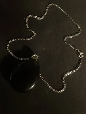 Silverhalsband med svart droppe - Snyggt halsband med en tunn silverfärgad kedja och ett stort, droppformat svart hänge. Hänget har en blank yta och ger ett stilrent intryck. Perfekt för dig som gillar minimalistiska smycken med en twist. OBS! Materialet är okänt, kan ej garantera att det är rostfritt.