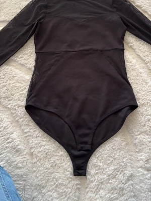 Svart meshbody från Amisu - Snygg svart body från Amisu med långa ärmar och meshdetaljer upptill. Tillverkad i ett stretchigt material som sitter tight och bekvämt. Perfekt att styla med jeans eller kjol för en trendig look.