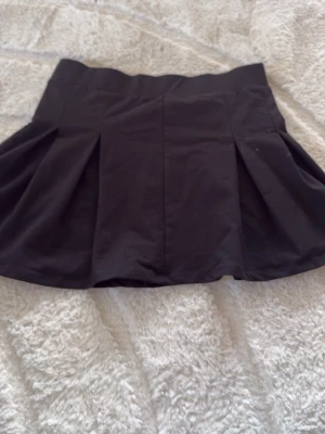 Svart kortkjol från 157 - Säljer en svart kortkjol från 157 med bred linning och snygga veck framtill. Kjolen har en klassisk, lätt utställd form som passar perfekt till många olika stilar. Enkel att matcha och superbekväm för både vardag och mer uppklädda tillfällen.