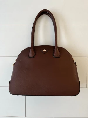 Brun handväska från Pauls Boutique - Stilren brun handväska från Pauls Boutique i konstläder med guldfärgad logga framtill. Väskan har två rejäla handtag och en rundad form, perfekt för dig som gillar klassisk och enkel design. Passar till många olika outfits och har en lyxig känsla.