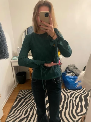 Grön ribbad tröja med knytband - Mörkgrön ribbad tröja med långa ärmar och dekorativa knytband vid ärmsluten. Tröjan har en figurnära passform och rund halsringning. Materialet ser mjukt och stretchigt ut, perfekt för en trendig och bekväm look.