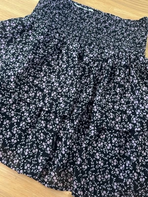 Svart blommig kortkjol med volang - Supersöt svart kortkjol med små ljusrosa blommor och volangdetaljer. Kjolen har en bred, smockad midja som ger en bekväm och snygg passform. Perfekt för dig som vill ha en luftig och trendig kjol till sommaren.