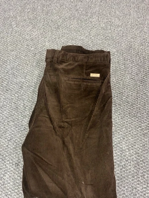 Bruna manchesterbyxor från Oscar Jacobson - Mörkbruna manchesterbyxor från Oscar Jacobson med klassisk rak passform och smala ribbor. Byxorna har bakficka med knapp och diskret logotypmärke ovanför fickan. Perfekta för dig som gillar retrostil och vill ha något bekvämt och snyggt.