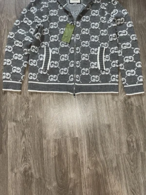 Grå ziptröja med GG-mönster från Gucci - Snygg grå ziptröja från Gucci med vitt GG-monogram över hela plagget. Tröjan har ribbade muddar och nederkant med vita detaljer samt hög krage. Tillverkad i mjukt stickat material som känns lyxigt och trendigt. Storlek S men passar xs också. Om du vill se mer bilder eller video då får du gärna skriva