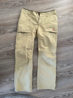 Beiga cargobyxor från Fjällräven - Snygga beiga cargobyxor från Fjällräven med raka ben och praktiska fickor på sidorna. Tillverkade i slitstarkt material och har den klassiska röda Fjällräven-loggan broderad på ena benet. Perfekta för dig som gillar outdoor och streetwear.