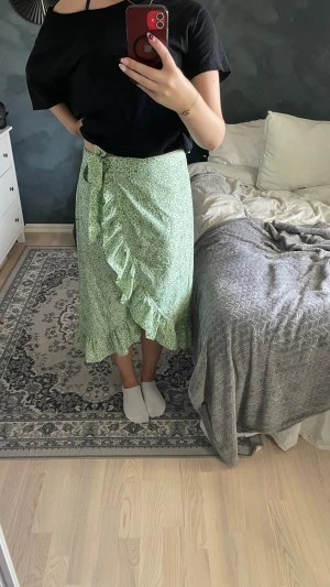 Grön blommig omlottkjol med volang - Säljer en ljusgrön omlottkjol med små blommor och volangkant. Kjolen har knyt i midjan och en flowy passform som ger en avslappnad och trendig look. Perfekt för vår och sommar!