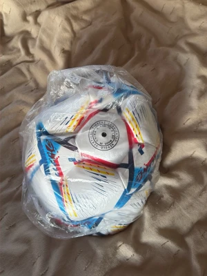 Vit fotboll med färgglada detaljer - Säljer en vit fotboll från Nivia med färgglada detaljer i blått, rött och gult. Bollen har ett modernt grafiskt mönster och är tillverkad i slitstarkt syntetmaterial. Perfekt för träning eller spontanspel med vänner.