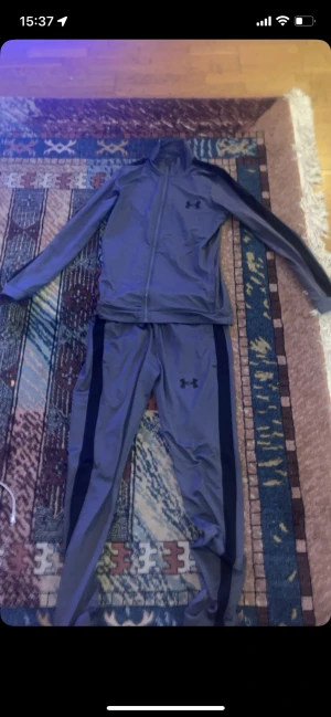 Grått träningsset från Under Armour - Säljer ett grått träningsset från Under Armour med svarta detaljer. Setet består av en zip-jacka och matchande byxor, båda med Under Armour-logga. Perfekt för gymmet eller löprundan. Materialet är troligtvis polyester för maximal rörelsefrihet.