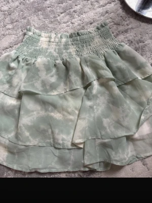 Ljusgrön volangkjol med resårmidja - Söt kortkjol i ljusgrönt med vitt mönster och bred resår i midjan. Kjolen har dubbla volanger och är gjord i ett tunt, luftigt material som ger en flowy känsla. Perfekt för vår och sommar!