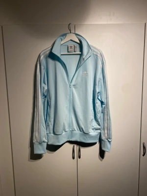 Ljusblå Adidas track jacket - Säljer en ljusblå Adidas track jacket med dragkedja framtill och klassiska vita ränder längs ärmarna. Jackan har hög krage, muddar vid ärmslut och nederkant samt två sidofickor. Perfekt för en sportig och avslappnad stil. Storlek M och true to size. Väldigt bra skick, sparsamt använd 