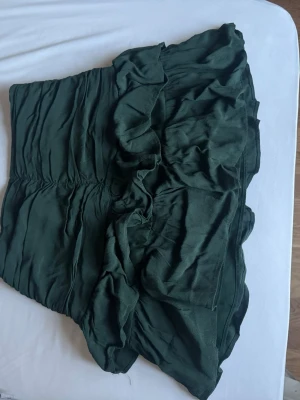 Mörkgrön volangkjol i polyester - Supersnygg mörkgrön kortkjol med volangdetaljer och rynkningar. Kjolen har en flowy känsla och är gjord i ett lätt, glansigt polyestertyg som ger en cool look. Perfekt att styla med en enkel topp eller stickad tröja.