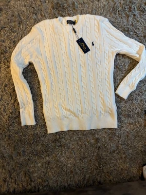 Vit kabelstickad tröja Polo Ralph Lauren - Kabelstickad vit tröja från Polo Ralph Lauren med rund halsringning och ribbade muddar. Tröjan har det klassiska mörkblå logobroderiet på bröstet och är tillverkad i mjukt material. Perfekt för dig som gillar stilren och tidlös design.