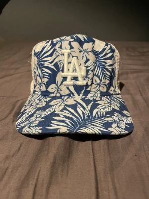 Blåvit LA keps med blommönster - Säljer en blåvit keps från New Era med LA-logga framtill. Kepsen har ett tropiskt blommönster och vit mesh baktill för extra ventilation. Justerbar snapback gör att den passar de flesta. Perfekt för dig som vill sticka ut med en unik stil.