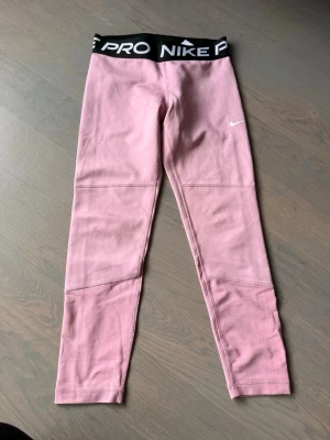 Rosa Nike Pro leggings - Snygga rosa Nike Pro leggings med svart elastisk midja och vit logga på låret. De har en tight passform och coola sömdetaljer på benen. Perfekta för träning eller chill. Materialet är stretchigt och bekvämt.