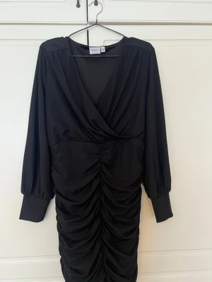 Svart omlottklänning från VILA - Snygg svart omlottklänning från VILA med långa ärmar och rynkade detaljer framtill. Klänningen har v-ringning och breda muddar vid ärmsluten. Perfekt för dig som gillar en stilren och trendig look.