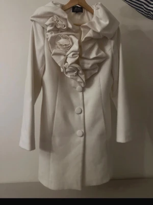 Vit rock med volang och rosetter - Snygg vit rock  med stora volanger och dekorativa tygrosor längs kragen. Rocken har knäppning framtill med stora tygklädda knappar och är figursydd för en elegant siluett. Perfekt för dig som vill sticka ut med en unik och trendig jacka.