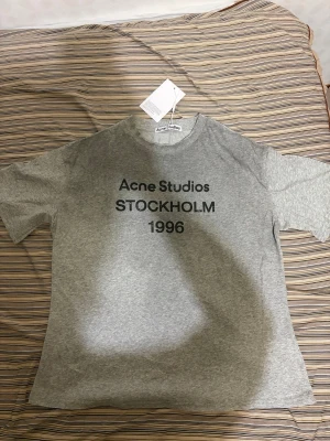 Acne studios t shirt  - Snygg grå t-shirt från Acne Studios med trycket 'Acne Studios STOCKHOLM 1996' på bröstet. Klassisk rund hals och korta ärmar. Perfekt för dig som gillar minimalistisk och stilren design. Materialet känns mjukt och skönt mot huden.