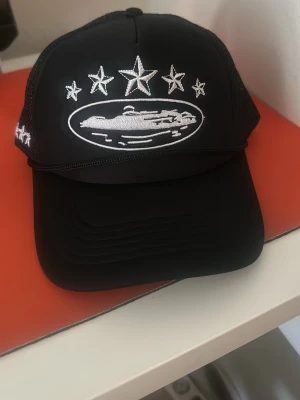 Svart meshkeps/ cortiez  - Säljer en svart keps från Corteiz med vit brodyr framtill som visar fem stjärnor och ett motiv, samt broderad logga bak. Kepsen har meshpaneler på sidorna och bak för extra ventilation och justerbar snapback. Perfekt för dig som gillar streetwear och vill sticka ut.