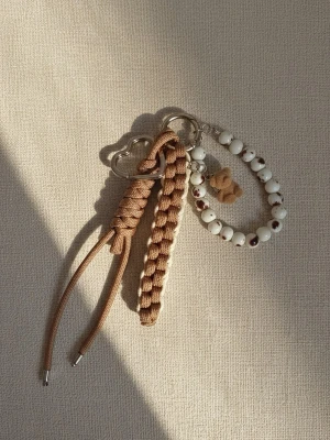 Handgjord Paracord Nyckelring & Väskcharm - Säljer en söt nyckelring med beige flätad rem i paracord, hjärtformat metallfäste och dekoration av vita pärlor med bruna detaljer samt en liten nallebjörn i filt. Perfekt att hänga på väskan eller använda till nycklar.
