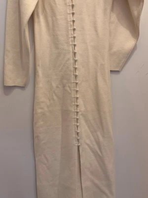 Krämvit långärmad klänning - Lång, krämvit klänning med raka ärmar och dekorativa knappar längs framsidan. Klänningen har en minimalistisk design och är tillverkad i ett mjukt, stickat material som ger en bekväm känsla. Perfekt för dig som gillar stilrena plagg med unika detaljer.
