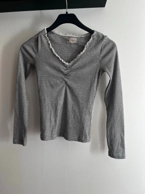 Randig långärmad topp Holly & Whyte - Säljer en svartvit, långärmad topp från Holly & Whyte. Toppen har smala ränder, v-ringning med söt volangkant och är figurnära i modellen. Perfekt för dig som gillar en enkel men ändå detaljrik stil. Älskar denhär tröjan men den har tyvärr blivit för liten över bysten