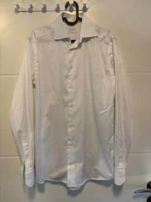 Vit skjorta från Eton - Klassisk vit skjorta från Eton med lång ärm och knappar framtill. Skjortan har en stilren design med bred krage och är tillverkad i bomull. Perfekt för en clean och uppklädd look. Hör av dig vid intresse!
